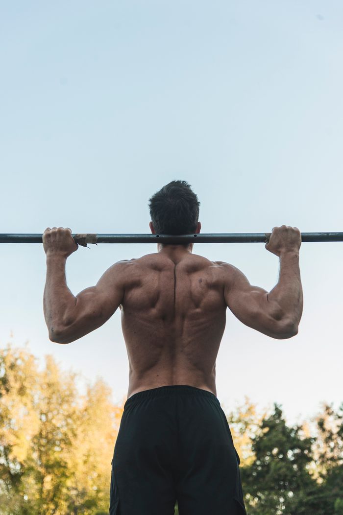 hero-img-01 Rear view of a strong man exercising shirtless with a barbell outdoors, showcasing strength and fitness.