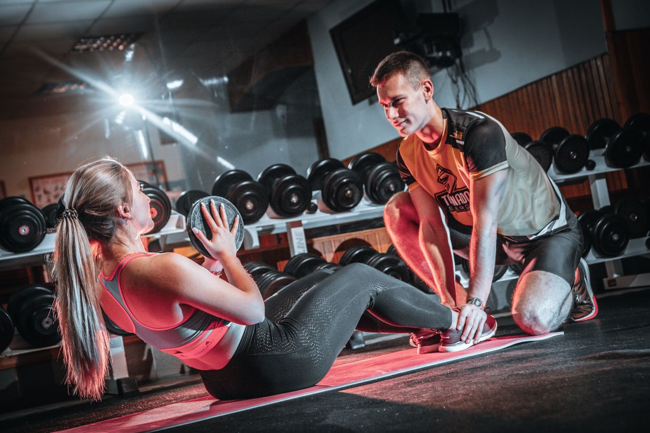 about-01 A personal trainer assists a woman doing sit-ups with a medicine ball in a gym setting.